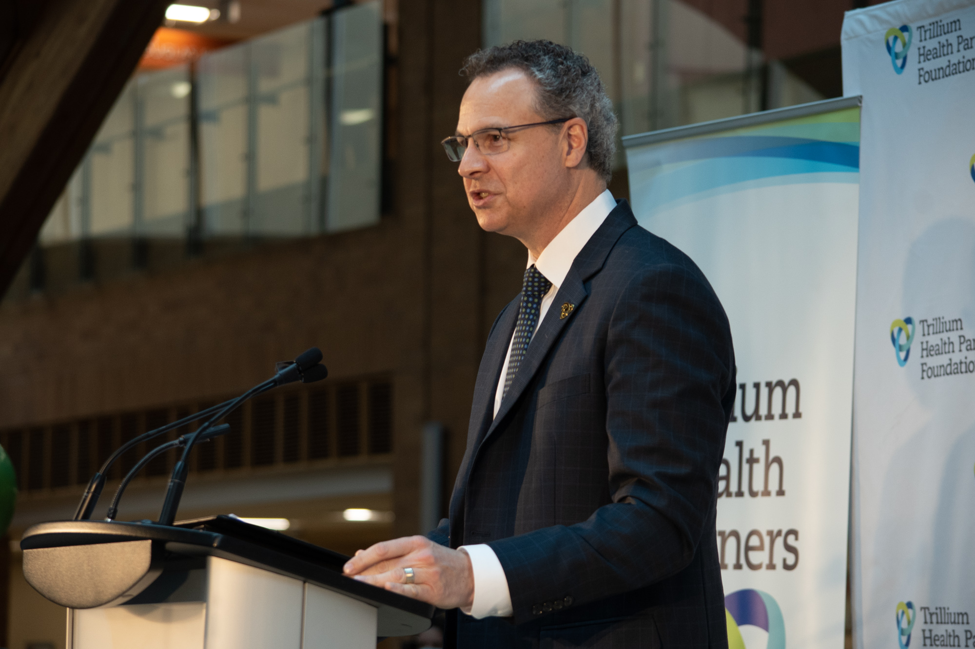 A man in a suit stands a podium in a large room with windows.