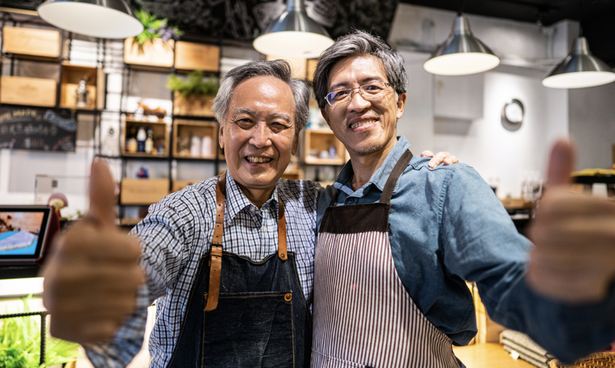 Two men wearing aprons look into the camera while giving the thumbs up sign in a grocery store.