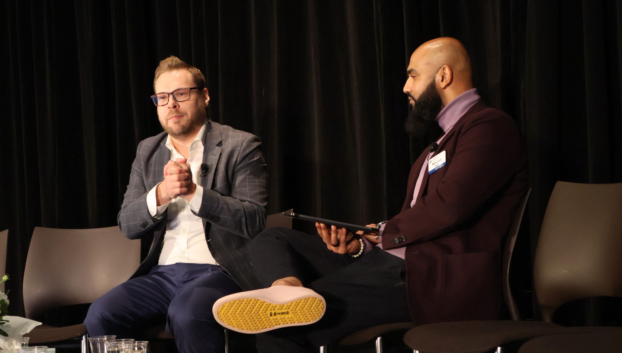 Two men in suits sit on a stage in chairs.