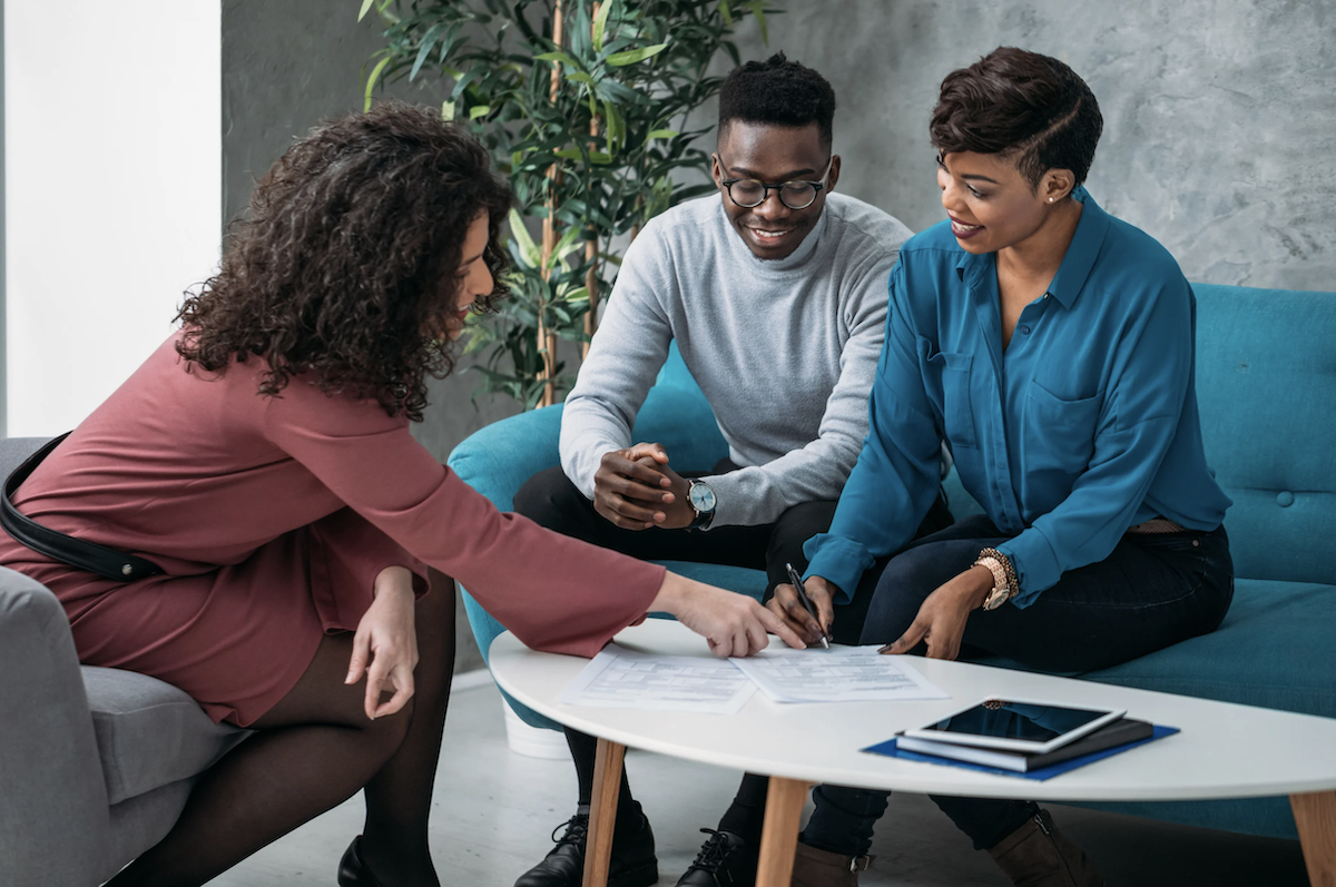 Three young, Black people wearing business casual sit around a low table looking at financial documents.