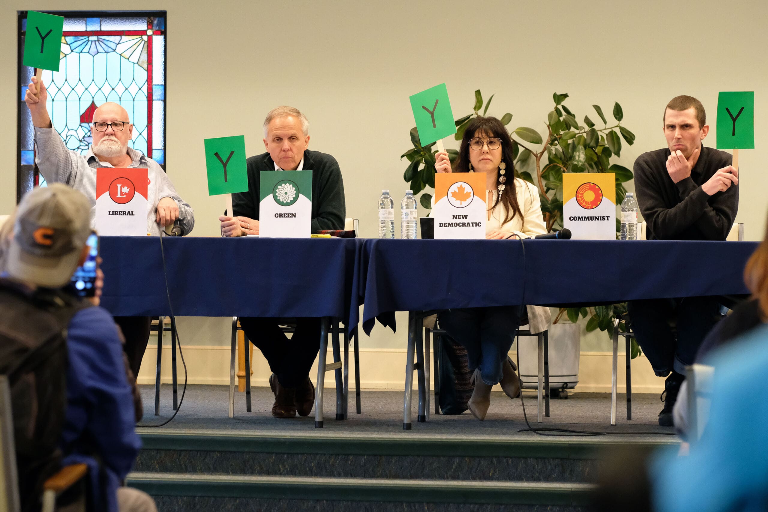 Ian MacIntyre of the Liberal Party, Gerry Gervais of the Green Party, Leah Gazan of the NDP and Cam Scott of the Communist Party take part in an election debate about accessibility at Winnipeg’s First Unitarian Universalist Church, while sitting behind a long table holding up green signs with the letter "Y" on them, on April 8, 2025