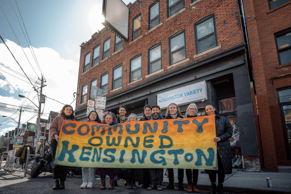 Members of the Kensington Market Land Trust stand in front of 54-56 Kensington Ave. in Toronto holding a multicoloured banner reading Community Owned Kensington. (Supplied photo/Kensington Market Land Trust)
