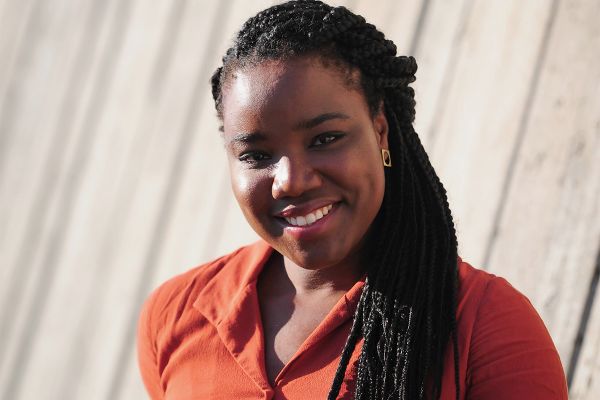 A Black woman in an orange shirt stands in-front of a concrete wall.